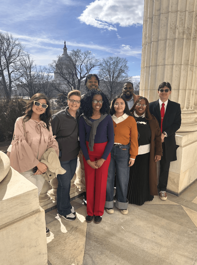A group of eight people pose together, smiling brightly at the camera as they stand in front of a classical column with the Capitol building on display in the background.