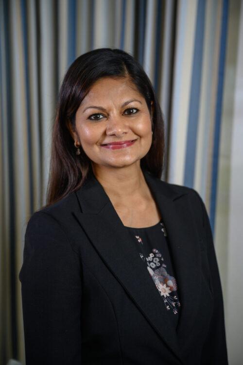 a portrait of Majabeen Rafiuddin in a black blazer and a dark floral shirt. She has dark hair and is standing in front of a striped blue and white curtain.